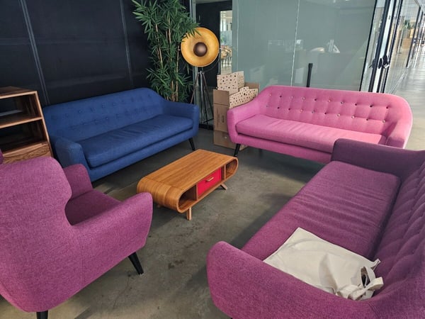 A photo of a brightly colored, mid-century modern style lounge area, featuring a blue sofa, a pink sofa, and two purple seating pieces arranged around a wooden coffee table with a red drawer.