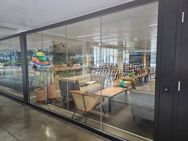 A photograph showing the interior of a modern office space through a large glass wall, featuring a lounge area with contemporary gray furniture, a coffee table, and potted plants.