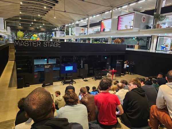 A high-angle photograph captures a large audience seated on tiered steps inside a modern, multi-level indoor venue, attending a presentation on a stage labeled "MASTER STAGE." The audience is primarily seen from behind, focused on a screen displaying content near the stage.