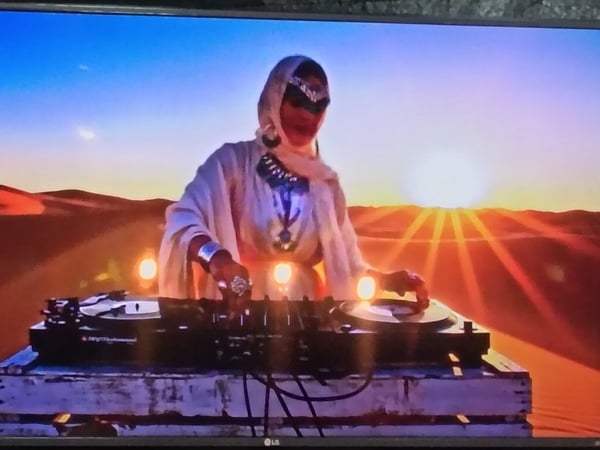 A person in traditional desert attire performs as a DJ at a turntable setup in a desert landscape during sunset.