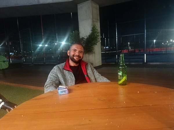 A man is sitting at a wooden table outdoors at night, smiling towards the camera with a beer bottle and a pack of cigarettes in front of him.