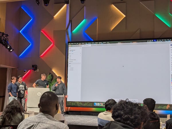 A photo capturing a group of young men presenting on a stage in an auditorium, standing in front of a large blank screen displaying a software interface.