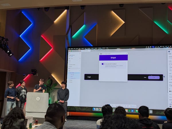 Four young men are presenting a software demonstration on a stage in a modern auditorium, showcasing a design assistant tool called 'Shipit' on a large projection screen. The stage background features striking, geometrically arranged acoustic panels illuminated by colorful neon-like LED strips.