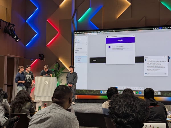A photograph capturing a software demonstration or presentation event, showing four young men on a stage in front of a large screen displaying a design tool interface (likely Figma) with an AI feedback pop-up labeled 'Shipit'. The backdrop features dramatic geometric wall paneling illuminated by colorful neon strip lights.