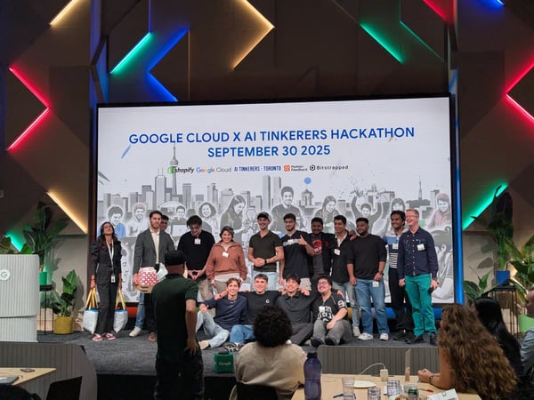 A group of young adults poses on a stage in front of a large screen advertising the 'Google Cloud x AI Tinkerers Hackathon' event, set in a modern indoor venue with colorful geometric lighting.