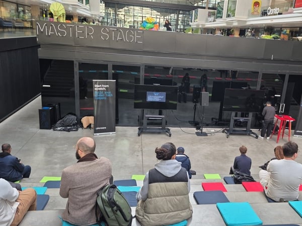 A photograph capturing an indoor event setting where an audience is seated on tiered steps, facing a stage labeled 'MASTER STAGE' in a large, modern atrium structure. The stage area features two large display screens and promotional banners for 'pioneers'.