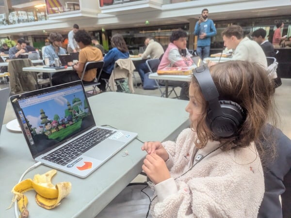 A young girl wearing black JBL headphones sits at a table, focused on a laptop screen displaying a colorful cartoon scene, while numerous people work on computers in a large, busy indoor atrium or event space in the background.