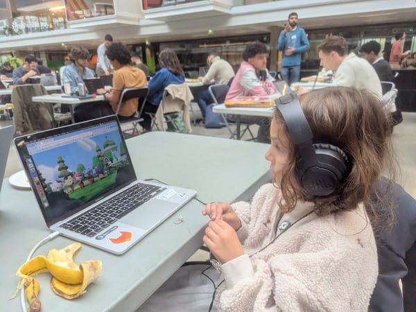 A candid photograph showing a young girl wearing black JBL headphones and a fluffy beige jacket, sitting at a table and looking at a laptop screen displaying a colorful cartoon scene, amidst a large group of people working in a busy indoor hall.