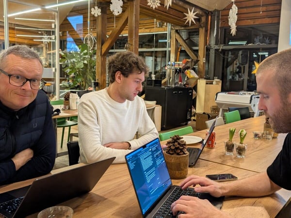 A photograph showing three men seated around a long wooden table in a rustic indoor workspace, working on open laptops during what appears to be a meeting or collaboration session.