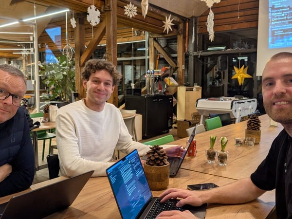 Three men are seated closely at a long wooden table with open laptops in a rustic indoor space, likely an office or co-working area, decorated with winter holiday ornaments.