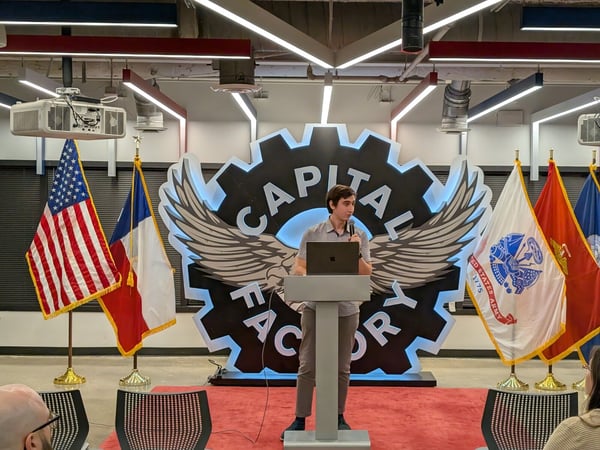A photograph showing a young man giving a presentation at a podium in an indoor event venue, standing in front of a large, illuminated backdrop featuring the 'CAPITAL FACTORY' logo, flanked by the US flag, the Texas flag, and military flags.