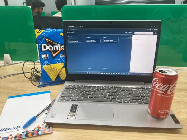 A silver laptop sits open on a wooden desk, displaying a Kanban board, flanked by a bag of Doritos and a can of Coca-Cola.