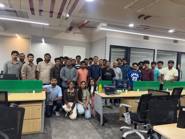 A large group of young professionals, primarily men, posing for a group photograph in a modern office environment with desks and computer equipment.