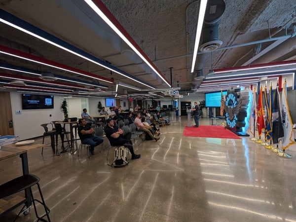 A man delivers a presentation to a seated audience in a modern, industrial-style event space featuring a large 'Capital Factory' logo and a row of military flags.