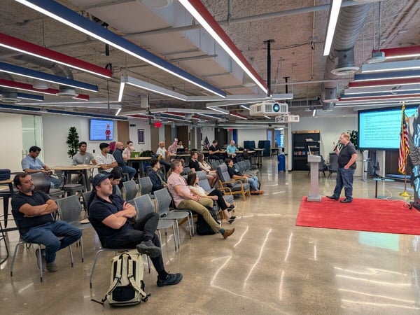A man stands on a small red carpet giving a presentation to a seated audience in a modern, industrial-style office space.