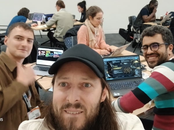 A selfie taken indoors showing three men in the foreground smiling at the camera, while several other participants work diligently on laptops at tables behind them in what appears to be a workshop or hackathon setting.