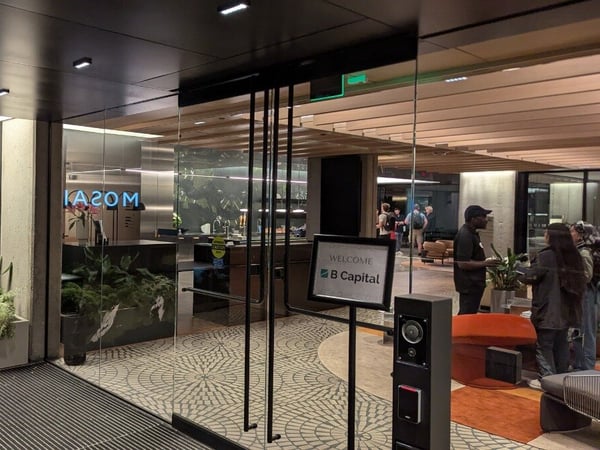 A photograph showing the modern entrance of an office building with a glowing 'MOSAIC' sign on the left wall and a glass door leading into a lobby where a 'WELCOME B Capital' sign is displayed.