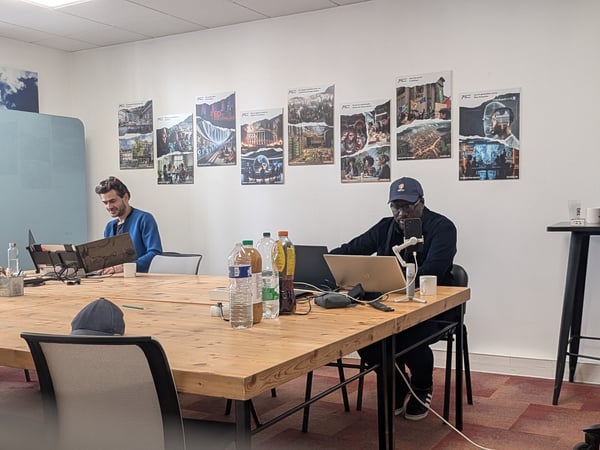 Two men are working on their laptops at a large wooden table in a bright office space decorated with several posters on the wall.