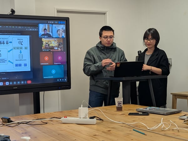 Two individuals are standing next to a large touchscreen display presenting a technical diagram for a project called 'Posey'. A laptop sits on a high table in front of them, and a power strip with cables is visible on the wooden table in the foreground.