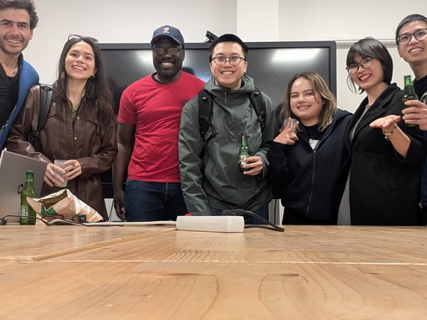 A group of seven diverse individuals are smiling and posing for a photo behind a large wooden table in an office or meeting room setting.