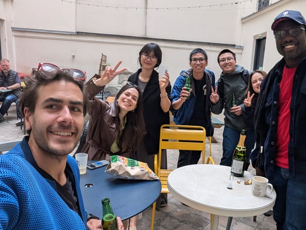A group of diverse friends or colleagues are smiling for a group selfie in an outdoor courtyard, enjoying drinks and snacks.