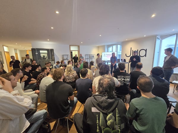 A large group of people gathered in a modern office space for a presentation, with speakers standing at the front near a screen and a large 'Jua' logo on the wall.