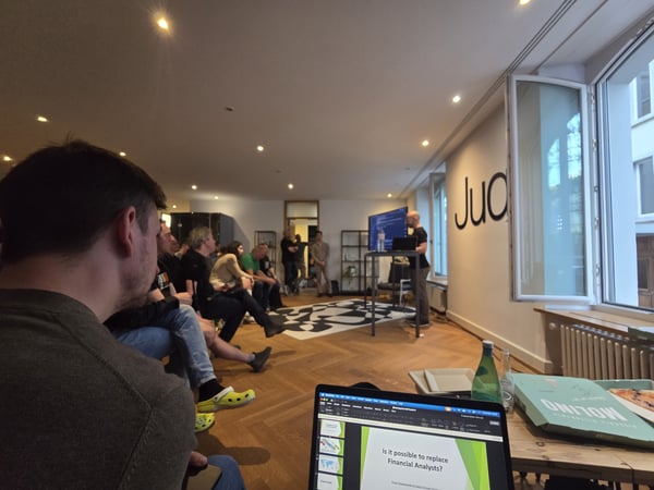 A group of people attending a presentation in a modern indoor space. A speaker stands at the front near a screen, while a laptop in the foreground displays a slide about financial analysts.