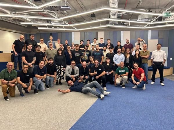 A large group of approximately 48 people, likely colleagues, pose for a group photo inside a modern office space featuring exposed ceilings and blue accent walls.