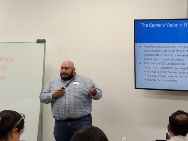 A candid photograph capturing a business presentation in a conference room, showing a bearded male presenter standing next to a whiteboard and a large screen displaying a slide about 'The Carver's Vision = The Business Use Case.' Several audience members are visible in the foreground with their backs to the camera.