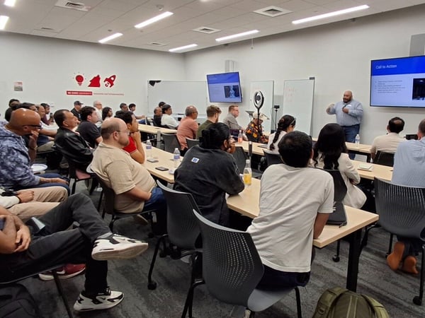 A photograph showing a large group of people attending a presentation or lecture in a modern conference room setting, with two presenters standing near dual display screens at the front of the room.