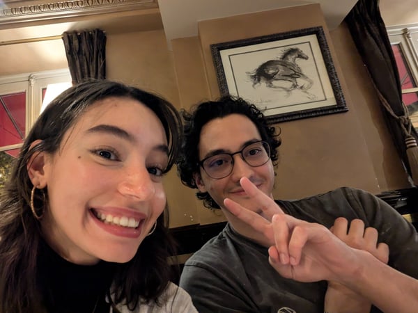 A close-up selfie shows a smiling young woman and a young man posing indoors in a warmly lit room, possibly a restaurant or bar.