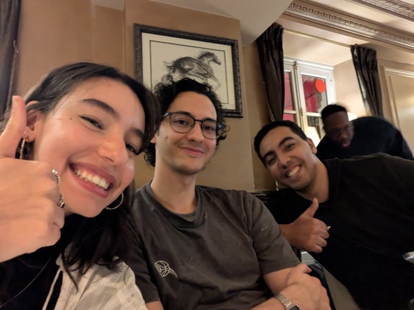 A group selfie taken indoors shows four people, with three in the foreground smiling and giving thumbs-up gestures, in what appears to be a warmly lit restaurant or ornate room.