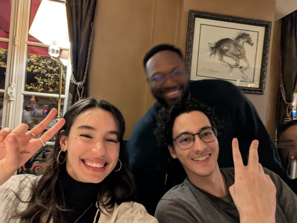 A close-up, candid photo shows three young people smiling and posing for the camera in a dimly lit restaurant setting, with the two foreground individuals making peace signs.