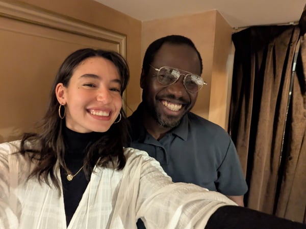 A close-up selfie shows a smiling woman and a man wearing glasses, both looking directly at the camera with wide smiles, likely taken indoors in a warmly lit setting.
