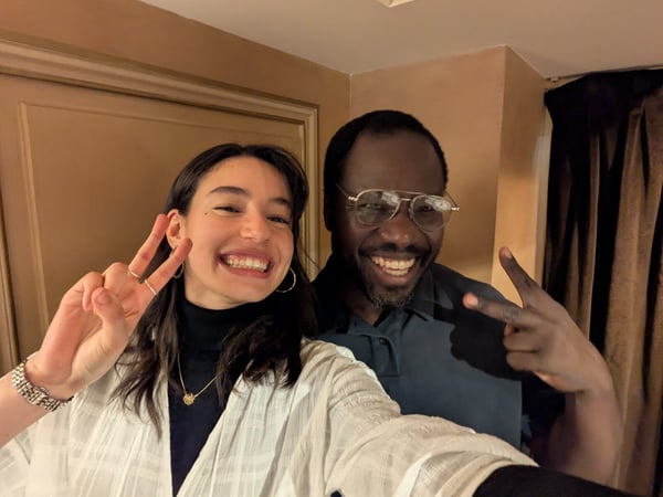 A close-up selfie shows a young woman and an older man smiling widely at the camera while both make peace signs with their hands, set indoors against a warm, tan background.