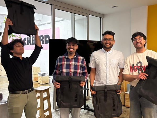 Four young men are posing indoors, possibly at a startup or product launch event, displaying black roll-top backpacks against a backdrop featuring a bright yellow accent wall and a partially visible banner.