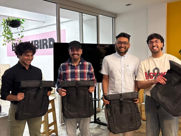 Four young men stand side-by-side indoors, smiling and posing while holding identical dark gray roll-top backpacks branded with "ElevenLab."
