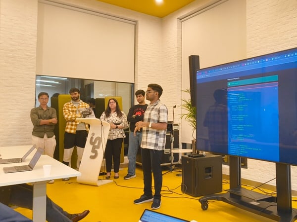 A group of five people are gathered in a brightly colored indoor space, where one individual is giving a presentation or demonstration of code shown on a large vertical monitor.