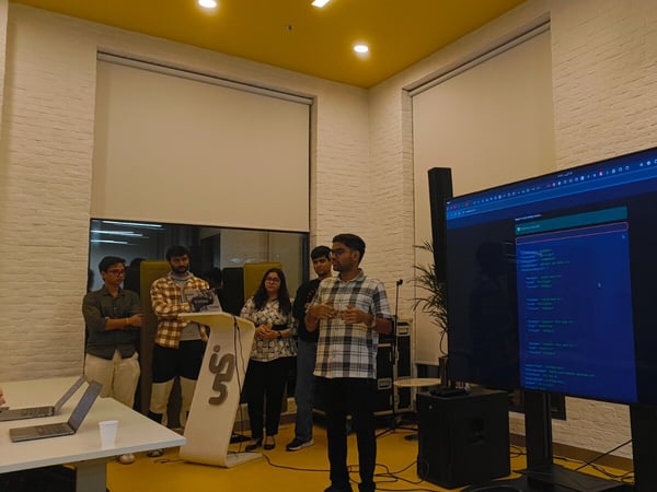 A group of five individuals is gathered indoors, where one man is giving a presentation or technical demonstration displayed on a large monitor showing code or data structures.