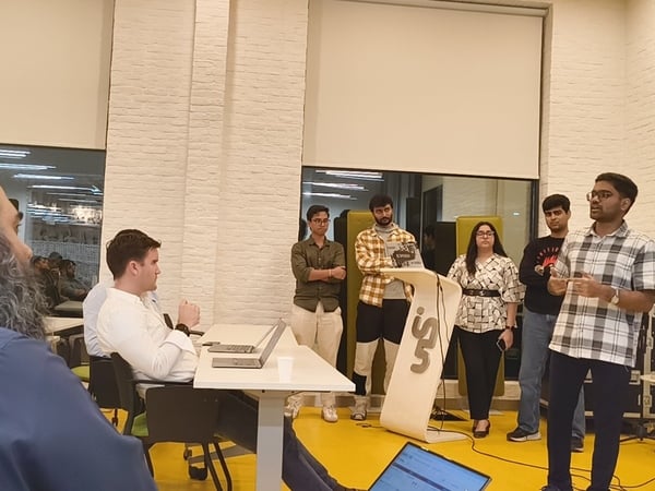 A candid photograph taken indoors during a presentation or meeting, showing a young man speaking near a stylized podium while several colleagues stand beside him and attendees observe from the foreground.