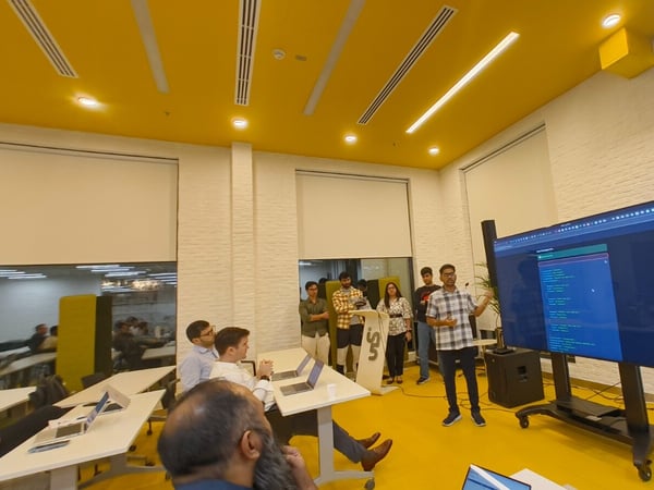 A man presents technical information, likely code, displayed on a large screen to an audience gathered in a modern, brightly lit room featuring yellow floors and ceilings.