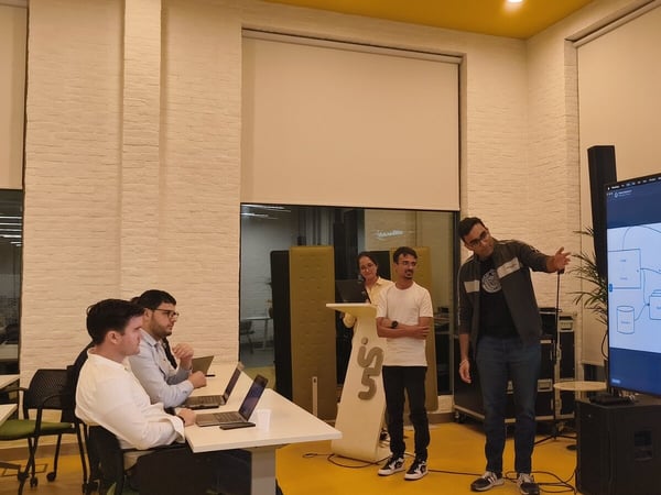 A photo capturing an indoor technical presentation where a man points to a complex flow diagram displayed on a large screen while several attendees observe, some using open laptops.