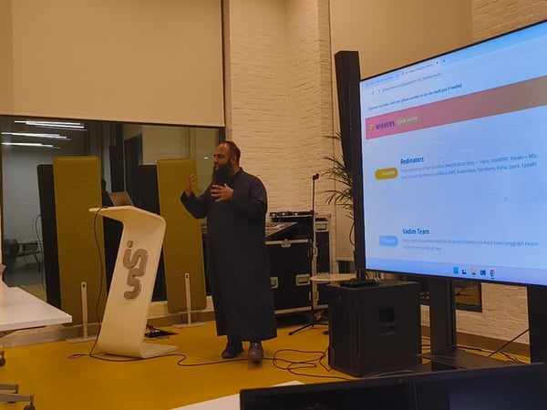 A man with a beard, dressed in a long dark robe, is giving a presentation or speech next to a modern white podium in an indoor venue with bright yellow flooring, while a large vertical screen displays a list of hackathon or competition winners and team details.