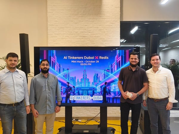 Four men are standing side-by-side in an indoor event space, posing in front of a large digital screen that displays promotional material for an AI Tinkerers Dubai and Redis mini hack event.