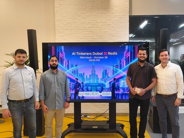Four men stand posing for a group photograph indoors in front of a large digital screen displaying promotional information for an AI Tinkerers Dubai and Redis hackathon event.