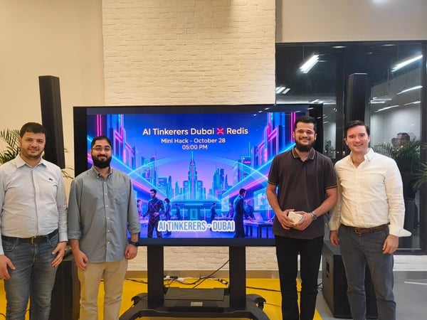 Four men stand side-by-side in an indoor event space, posing in front of a large digital screen that displays promotional graphics for an 'AI Tinkerers Dubai X Redis Mini Hack' event.