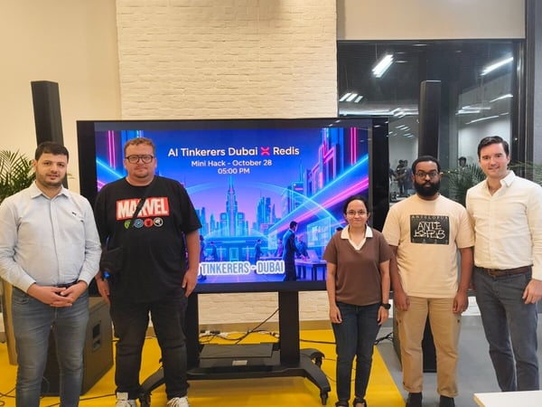 A group of five people (four men and one woman) are posing for a photo in front of a large digital screen at an indoor event, which displays branding for an "AI Tinkerers Dubai X Redis Mini Hack."