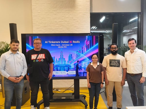 Five individuals, four men and one woman, stand side-by-side posing for a photograph in front of a large digital screen advertising an 'AI Tinkerers Dubai X Redis Mini Hack' event.