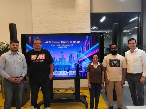 A group photo featuring five individuals (four men and one woman) standing indoors in front of a large digital screen displaying information for an event titled 'AI Tinkerers Dubai X Redis Mini Hack'.