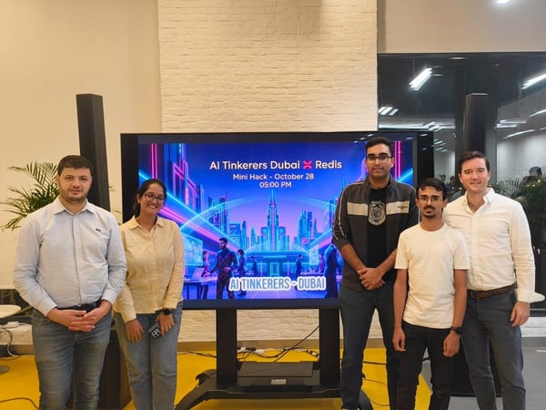A group of five people, consisting of four men and one woman, pose together in an indoor event space in front of a large digital screen advertising an 'AI Tinkerers Dubai X Redis Mini Hack.'